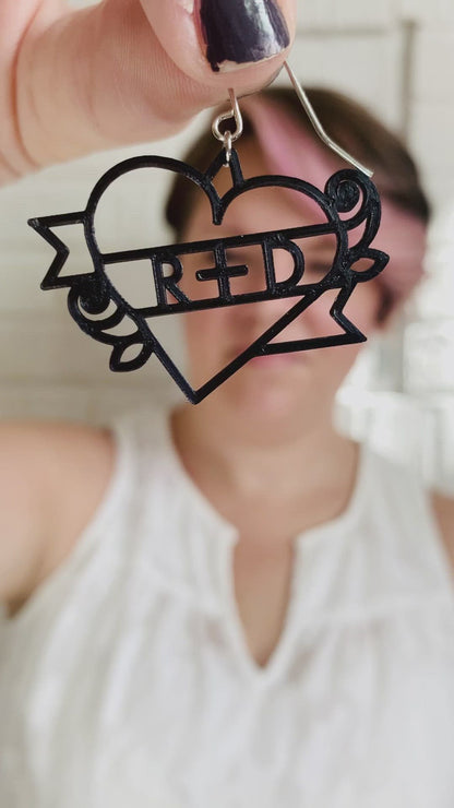 This is a video of Rebekah Thornhill, owner of R+D, showing a 3D printed earring. First she shows the earring close up. It is a bright red color and shaped as a heart with a banner twisting around it with roses on the sides. Writted on the banner is R+D. After showing the earring, she puts it on and shows the size of the earrings when being worn.