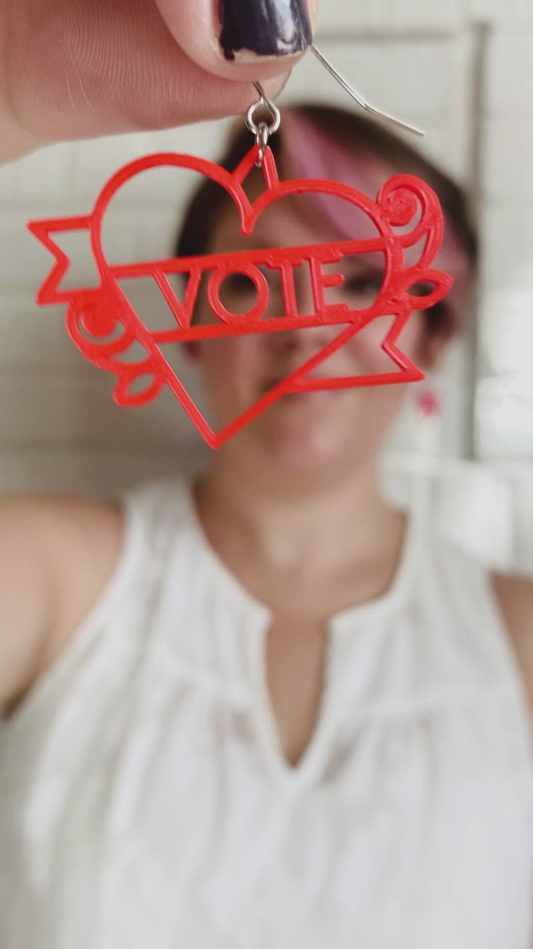 This is a video of Rebekah Thornhill, owner of R+D, showing a 3D printed earring. First she shows the earring close up. It is a bright red color and shaped as a heart with a banner twisting around it with roses on the sides. Writted on the banner is VOTE. After showing the earring, she puts it on and shows the size of the earrings when being worn. 