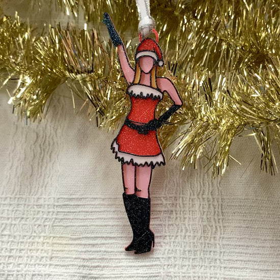 There is a white fabric background with gold garland. Laying on top of these is a 3D printed ornament from R+D. The ornament is printed in a plant based filament. It is shaped like Regina George from the movie Mean Girls. She is striking the iconic pose at the beginning of performing Jingle Bell Rock, wearing black gloves and boots and a red outfit with white trim. The entire ornament is covered in glitter to shimmer and shine in the light.