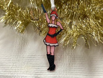 There is a white fabric background with gold garland. Laying on top of these is a 3D printed ornament from R+D. The ornament is printed in a plant based filament. It is shaped like Regina George from the movie Mean Girls. She is striking the iconic pose at the beginning of performing Jingle Bell Rock, wearing black gloves and boots and a red outfit with white trim. The entire ornament is covered in glitter to shimmer and shine in the light.