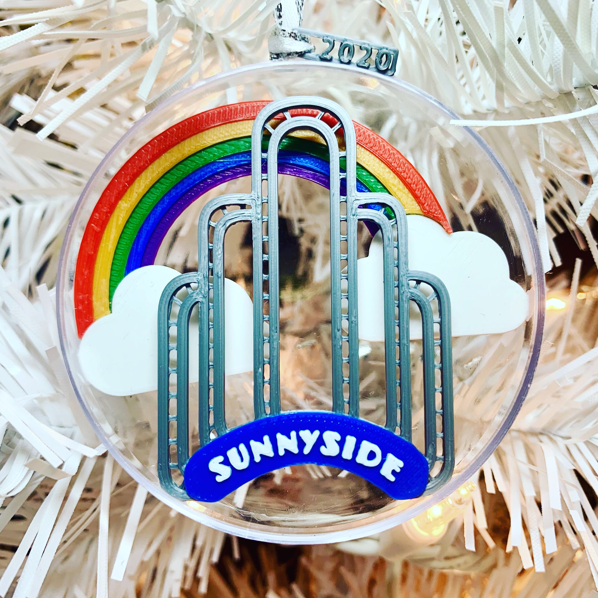 An image of our limited edition ornament for 2020--It features the art deco arch in Sunnyside, Queens, NYC, with a rainbow and clouds behind it. When you flip it over, there is a small fairy door at the borrow of the arch. 