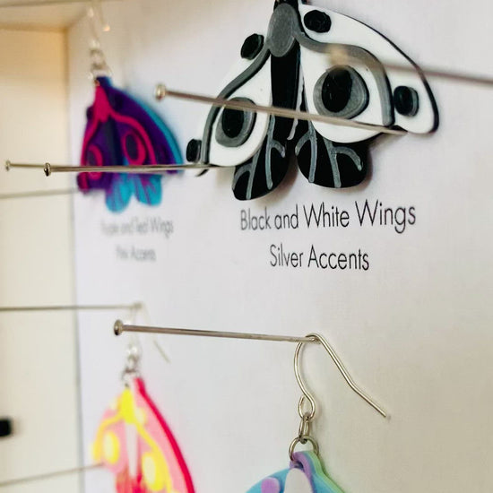 Shown in a white shadow box are six different colors of R+D 3D printed moth earrings. They are each held in place with pins like a shadow box in a natural history museum. The six colors are labeled: Purple and Teal Wings, Pink Accents; Black and White Wings, Silver Accents; Purple and Navy Wings, Blue Accents; Hot Pink and Light Pink Wings, Yellow Accents; Mint and Light Blue Wings, White Accents; and White and Light Blue Wings, Gold Accents.