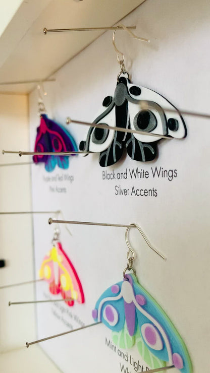 Shown in a white shadow box are six different colors of R+D 3D printed moth earrings. They are each held in place with pins like a shadow box in a natural history museum. The six colors are labeled: Purple and Teal Wings, Pink Accents; Black and White Wings, Silver Accents; Purple and Navy Wings, Blue Accents; Hot Pink and Light Pink Wings, Yellow Accents; Mint and Light Blue Wings, White Accents; and White and Light Blue Wings, Gold Accents.