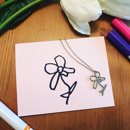 In the middle of the frame is a pink card with a flower drawn on it. Next to the card is a necklace with a 3D printed pendant that is the same as the  drawing.