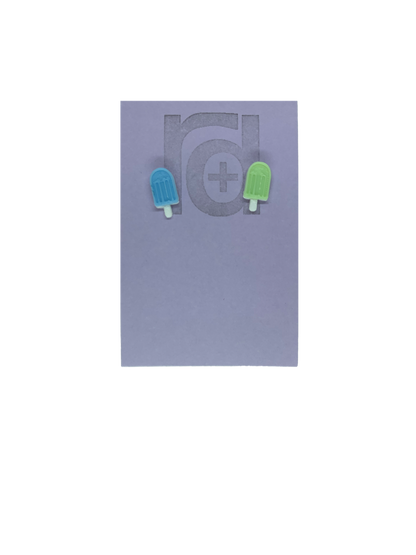 Two small and detailed R+D earrings are shown on a purple earring card. They are shaped as popsicles with white sticks. The earrings are mismatched, one popsicle is light blue and one is light mint green.