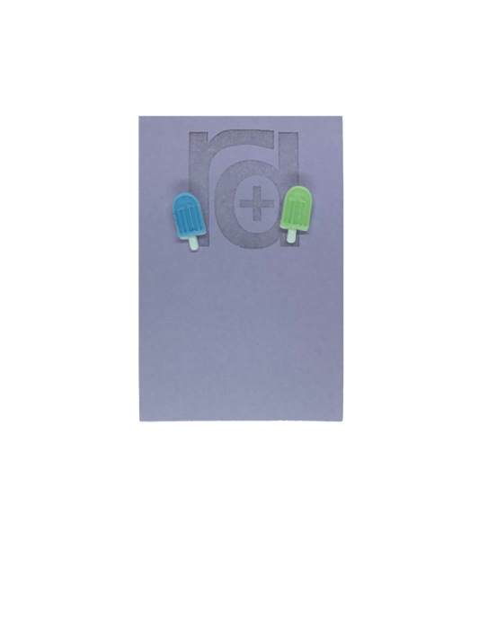 Two small and detailed R+D earrings are shown on a purple earring card. They are shaped as popsicles with white sticks. The earrings are mismatched, one popsicle is light blue and one is light mint green.