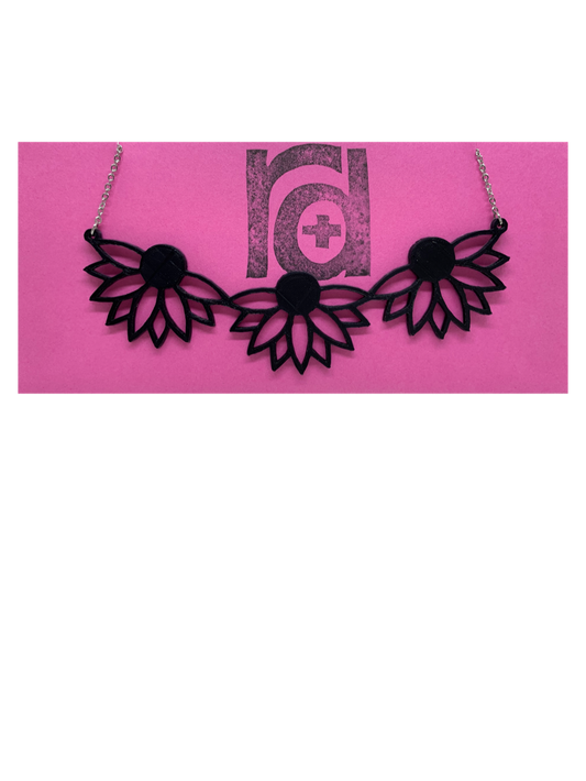 On a bight pink R+D necklace card is a 3D printed necklace. It is a bib necklace that stretches to have three daisys with petals stretching downward. This necklace is printed in a sustainable black filament. 