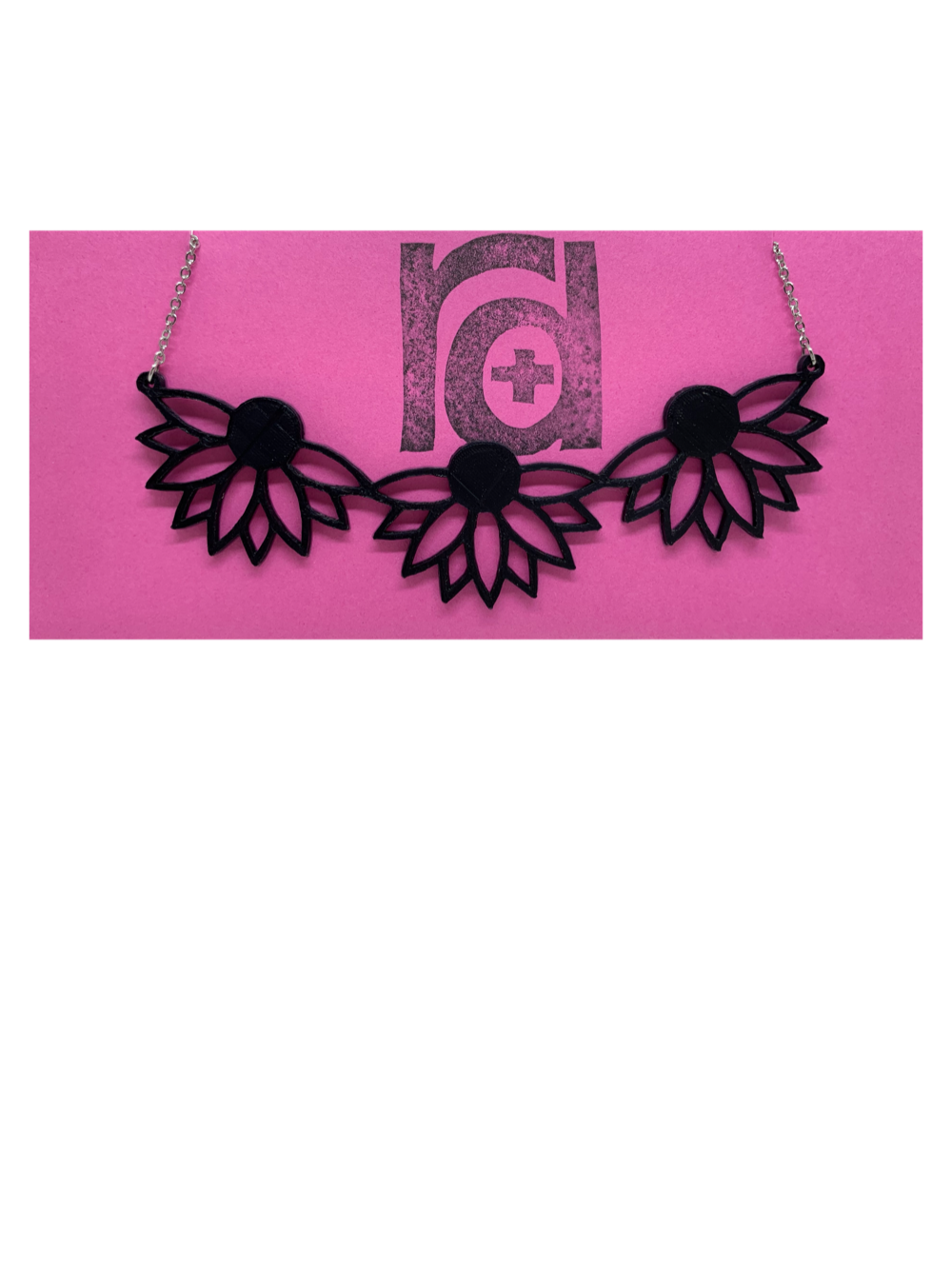 On a bight pink R+D necklace card is a 3D printed necklace. It is a bib necklace that stretches to have three daisys with petals stretching downward. This necklace is printed in a sustainable black filament. 
