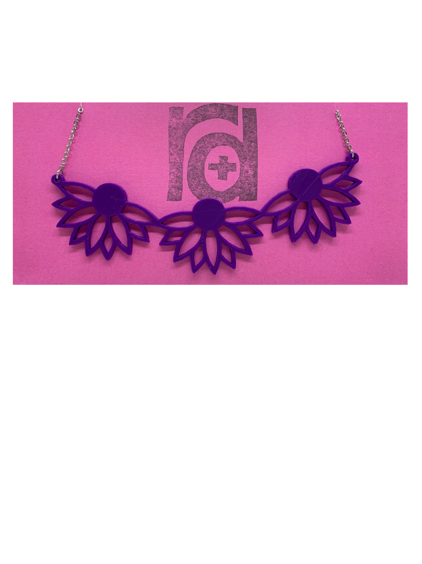On a bright pink R+D necklace card is a 3D printed necklace. It is a bib necklace that stretches to have three daisys with petals stretching downward. This necklace is printed in a sustainable purple filament. 