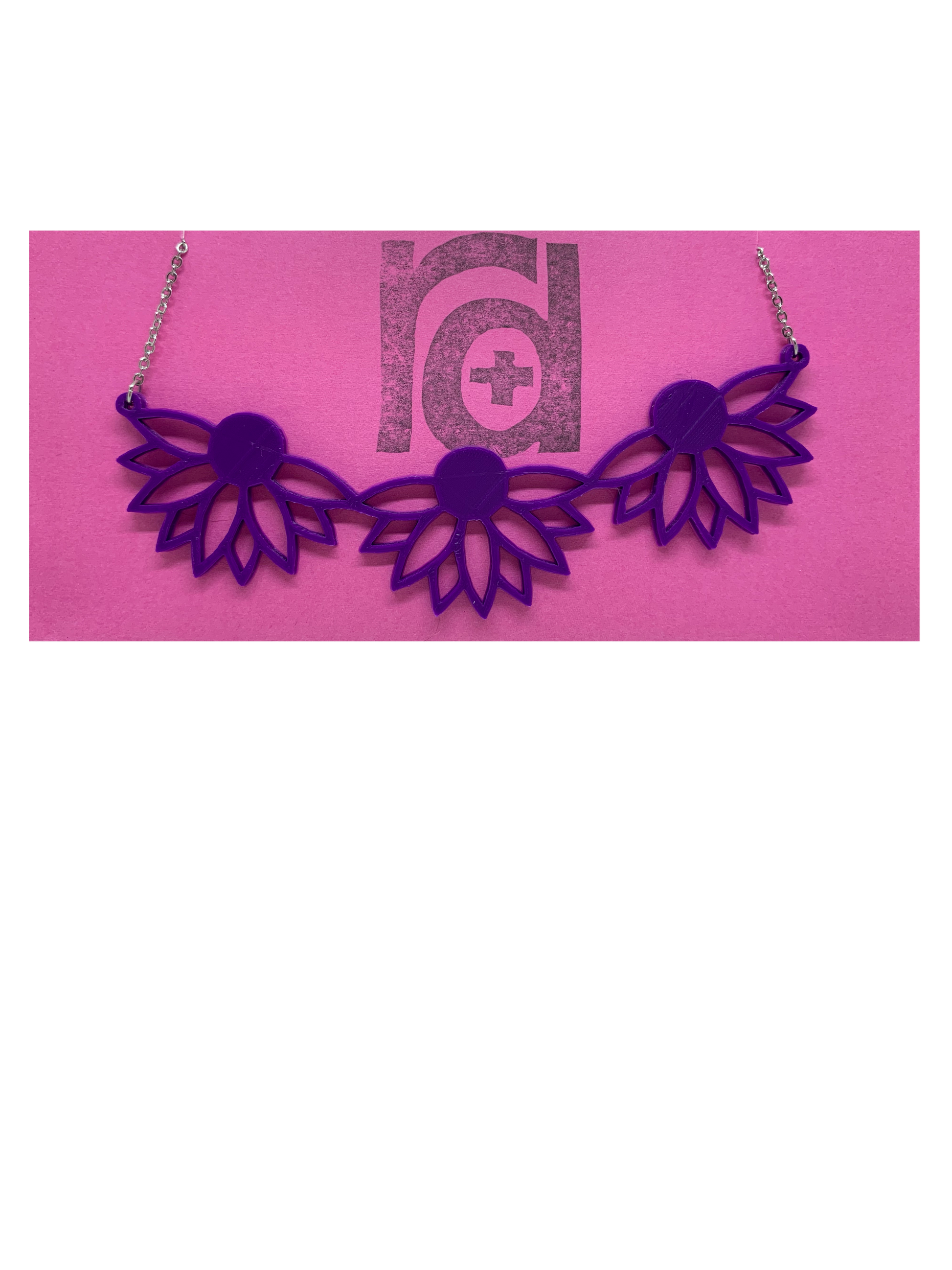 On a bright pink R+D necklace card is a 3D printed necklace. It is a bib necklace that stretches to have three daisys with petals stretching downward. This necklace is printed in a sustainable purple filament. 