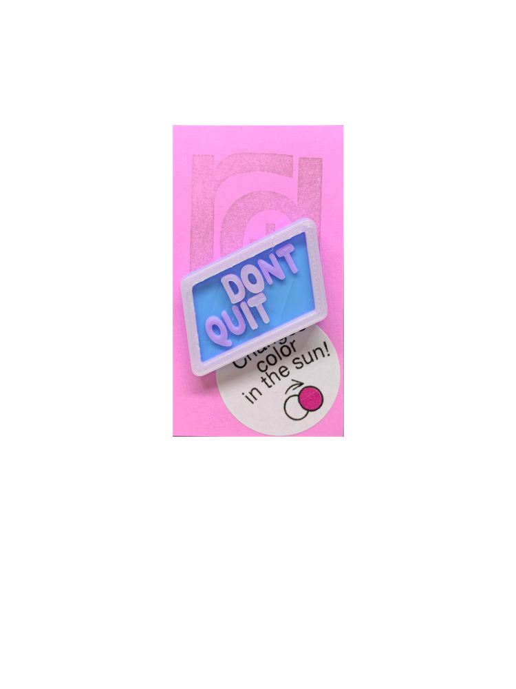 Shown on a pinkish lilac R+D card is 3D printed pin. It is in a light blue hexagon shape with purple writing that says DONT QUIT. There is also a opaque white layer on the pin. That layer is UV reactive and makes a border and reveals a hidden message in direct sunlight. Here it is shown out of the sunlight. 