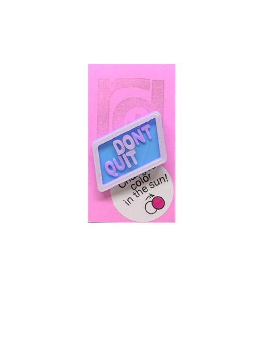 Shown on a pinkish lilac R+D card is 3D printed pin. It is in a light blue hexagon shape with purple writing that says DONT QUIT. There is also a opaque white layer on the pin. That layer is UV reactive and makes a border and reveals a hidden message in direct sunlight. Here it is shown out of the sunlight. 