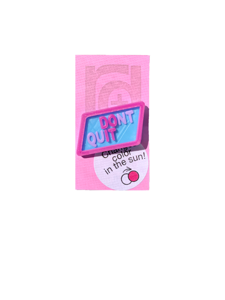Shown on a pinkish lilac R+D card is 3D printed pin. It is in a light blue hexagon shape with purple writing that says DONT QUIT. There is also a layer is UV reactive and makes a border and reveals a hidden message in direct sunlight. Here it is shown in direct sunlight, so you can see the words DO IT highlighted in bright hot pink.  
