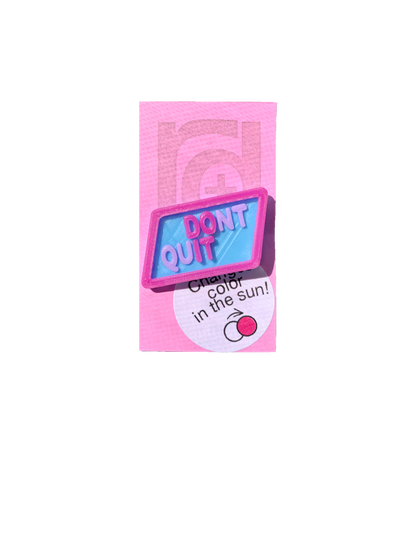 Shown on a pinkish lilac R+D card is 3D printed pin. It is in a light blue hexagon shape with purple writing that says DONT QUIT. There is also a layer is UV reactive and makes a border and reveals a hidden message in direct sunlight. Here it is shown in direct sunlight, so you can see the words DO IT highlighted in bright hot pink.  
