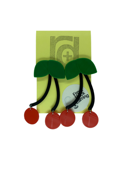 Hanging off of a yellow earring card are two statement earrings  shaped like cherries. They have green leaves that are worn on the front of the ear lobe with two black stems that lead to bright red cherries. The stem is worn behind the ear to give depth to this classic style that will match any picnic or rock a billy outfit.