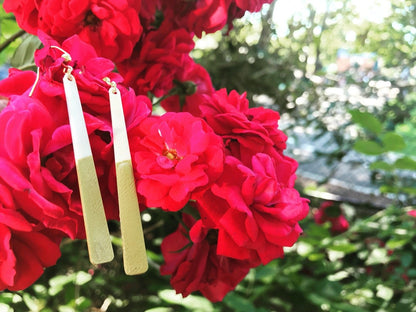 Hanging off of a cluster of bright red roses are two earrings. They are long white dangles that have been dipped in a gold metallic paint. 