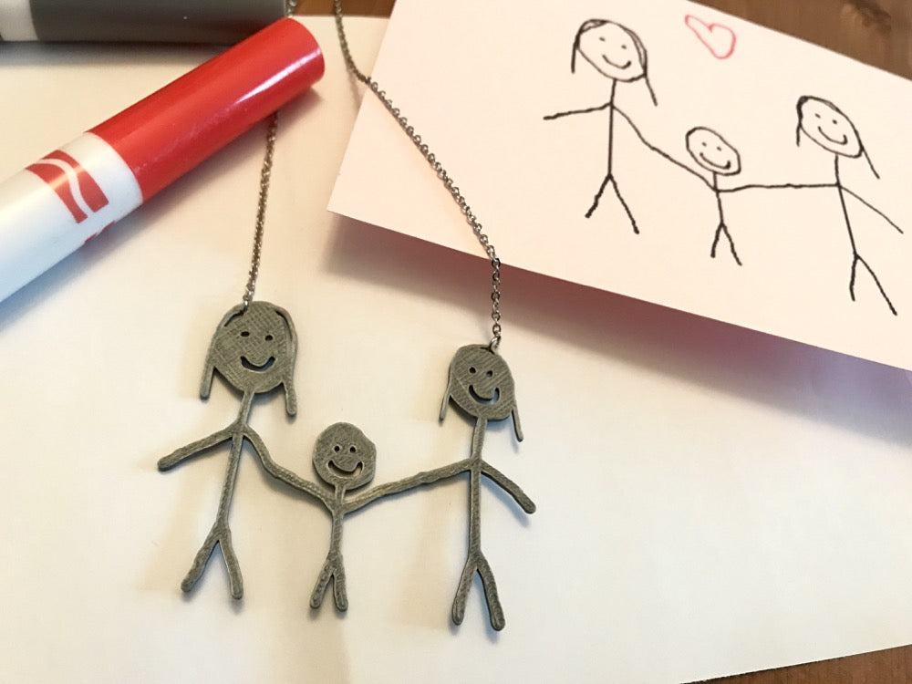 IN the background is a card with a stick figure family drawn on it. In the foreground is a necklace that features a 3D Printed pendant that is made from the drawing on the card. 