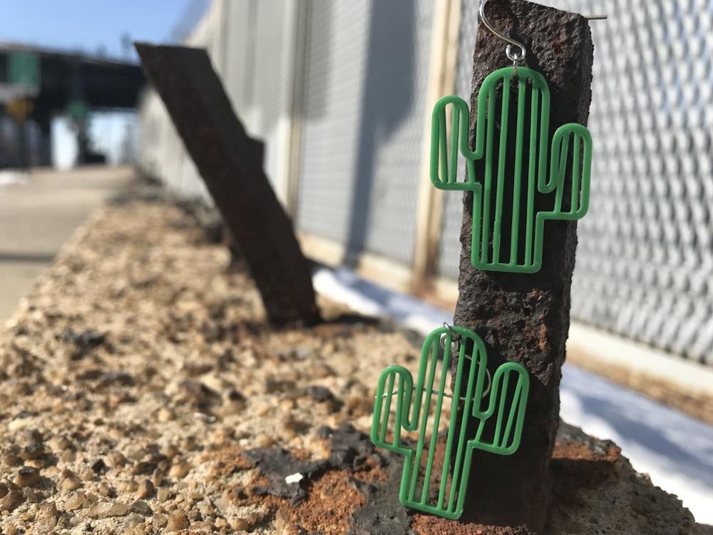 On a rocky curb with a blurred background are two earrings, each shaped like cactus. They each have two arms reaching out and vertical stripes to create texture. They are 3D printed in a green plant based material. 