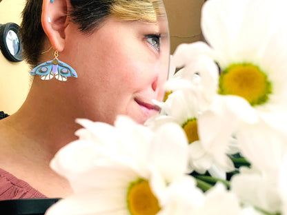 In the foreground is a bouquet of daisy flowers out of focus. Further away, but in focus, is a mirror reflecting a person wearing a 3D Printed earring from R+D. The earring is a moth that has white lower wings, light blue larger wings, a light purple body and gold details on the wings and body.