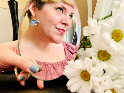 In the foreground is a bouquet of daisy flowers out of focus. Further away, but in focus, is a mirror being held up and reflecting a person wearing a 3D Printed earring from R+D. The earring is a moth that has white lower wings, light blue larger wings, a light purple body and gold details on the wings and body.