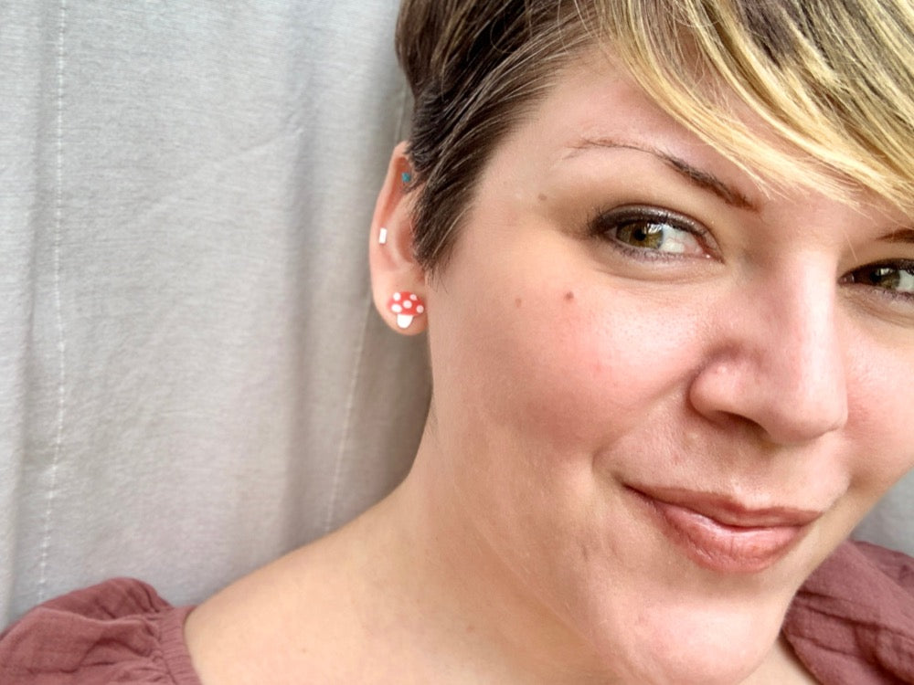 Smiling and looking past the camera is a woman wearing R+D 3D printed earrings. The tiny studs are small mushrooms with white stems, red caps, and white dots. 