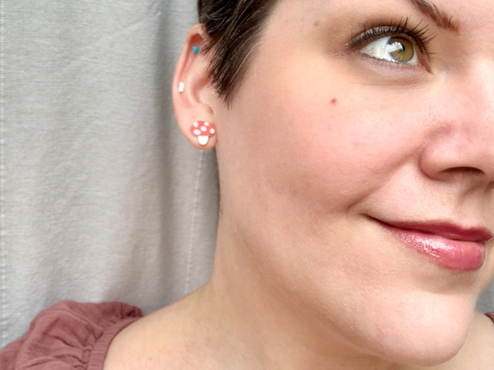 Smiling and looking up past the camera is a woman wearing R+D 3D printed earrings. The tiny studs are small mushrooms with white stems, red caps, and white dots.