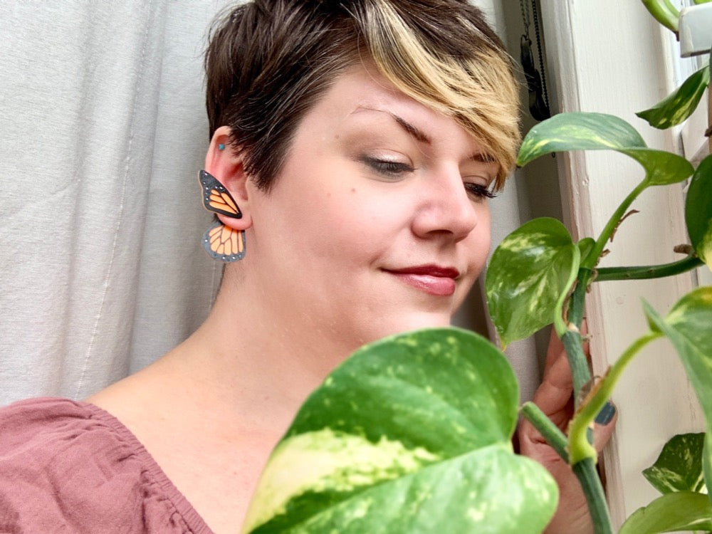 Standing by a window with climbing vines, a woman with short hair wears 3D Printed butterfly earrings. They are orange like monarch butterflies. The smaller top wing is worn in front of the earlobe and the larger wing hangs behind the earlobe. 