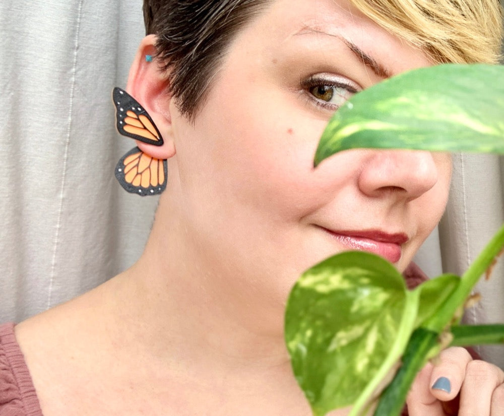 Standing by a window with climbing vines, a woman with short hair wears 3D Printed butterfly earrings. They are orange like monarch butterflies. The smaller top wing is worn in front of the earlobe and the larger wing hangs behind the earlobe.