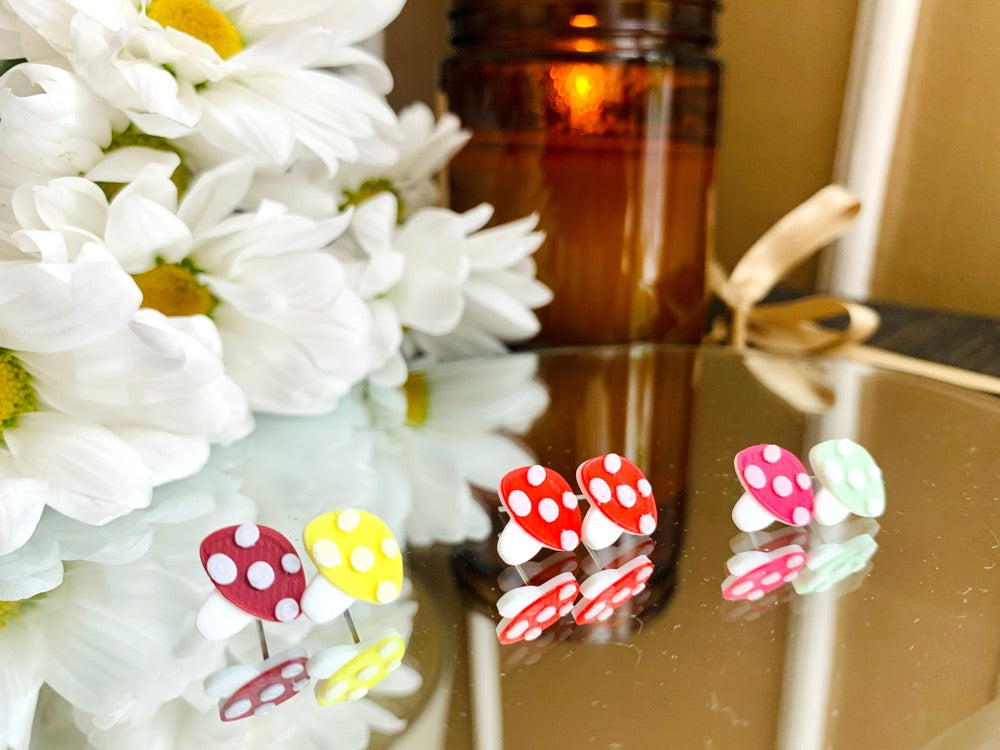 Resting on a mirror in front of a bouquet of daisies and a dark brown candle are three pairs of 3D printed R+D earring studs. The earrings are mushroom shapes with white stems and white dots on the caps. The first pair is merlot and yellow mismatched studs. The second is two red mushrooms. The final is hot pink and mint mushroom caps. 