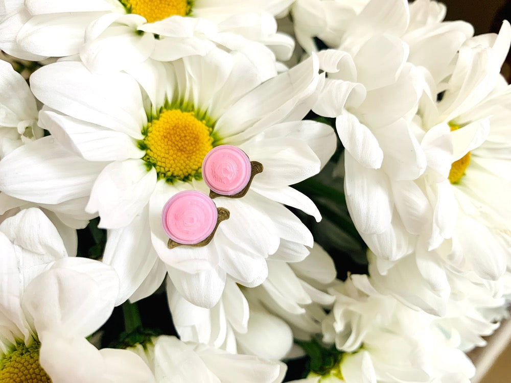 Resting in a bouquet of daisy flowers are two 3D printed R+D earrings. They are shaped like small snails with gold bodies and light pink swirled shells on their back. 