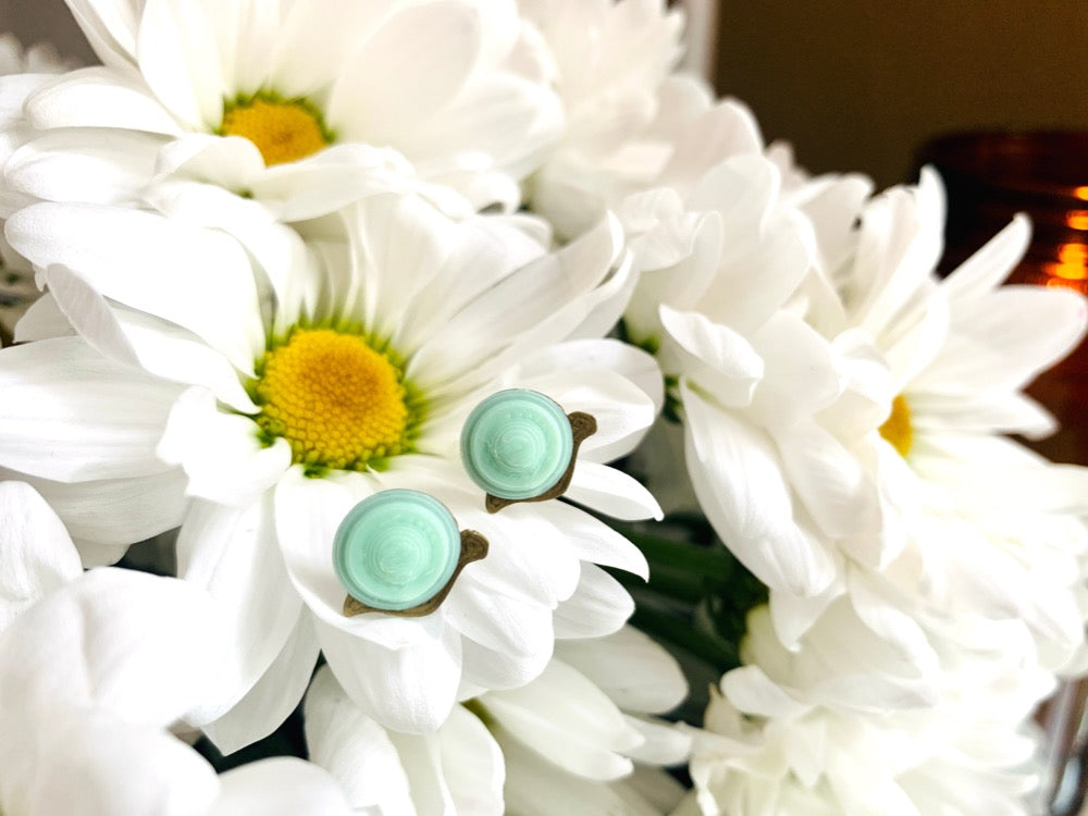 Resting in a bouquet of daisy flowers are two 3D printed R+D earrings. They are shaped like small snails with gold bodies and light mint green swirled shells on their back.
