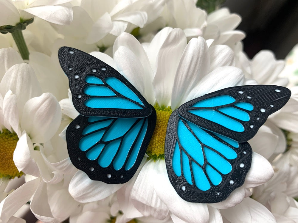 Resting on a bouquet of daisies is a pair of front back earrings that look like a bright blue and black monarch butterfly. These R+D earrings are 3D printed and have a small wing that is worn on the front of the earlobe and a larger wing that rests behind the earlobe.