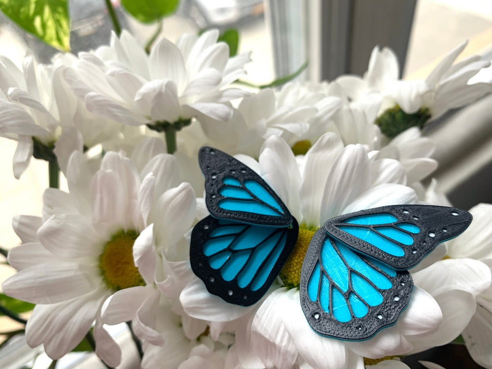 Resting on a bouquet of daisies is a pair of front back earrings that look like a bright blue and black monarch butterfly. These R+D earrings are 3D printed and have a small wing that is worn on the front of the earlobe and a larger wing that rests behind the earlobe.