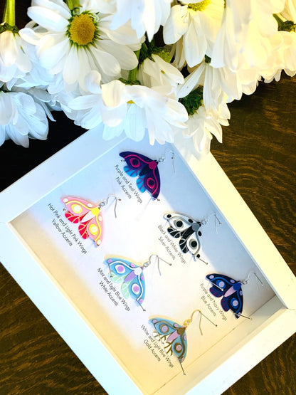 Shown in a white shadow box are six different colors of R+D 3D printed moth earrings. They are each held in place with pins like a shadow box in a natural history museum. The six colors are labeled: Purple and Teal Wings, Pink Accents; Black and White Wings, Silver Accents; Purple and Navy Wings, Blue Accents; Hot Pink and Light Pink Wings, Yellow Accents; Mint and Light Blue Wings, White Accents; and White and Light Blue Wings, Gold Accents. 
