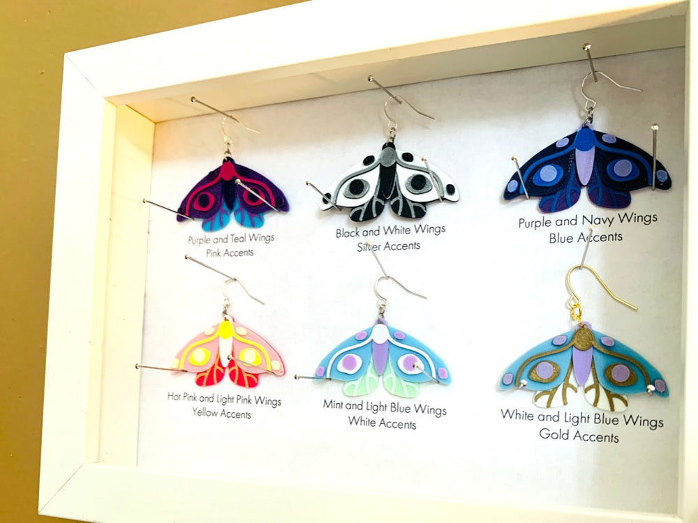 Shown in a white shadow box are six different colors of R+D 3D printed moth earrings. They are each held in place with pins like a shadow box in a natural history museum. The six colors are labeled: Purple and Teal Wings, Pink Accents; Black and White Wings, Silver Accents; Purple and Navy Wings, Blue Accents; Hot Pink and Light Pink Wings, Yellow Accents; Mint and Light Blue Wings, White Accents; and White and Light Blue Wings, Gold Accents.