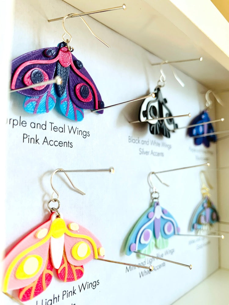 Shown in a white shadow box are six different colors of R+D 3D printed moth earrings. They are each held in place with pins like a shadow box in a natural history museum. The six colors are labeled: Purple and Teal Wings, Pink Accents; Black and White Wings, Silver Accents; Purple and Navy Wings, Blue Accents; Hot Pink and Light Pink Wings, Yellow Accents; Mint and Light Blue Wings, White Accents; and White and Light Blue Wings, Gold Accents.