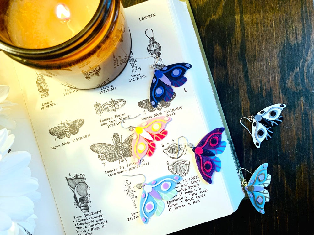 Shown on a old book with drawings are six different colors of R+D 3D printed moth earrings. The six colors are Purple and Teal Wings, Pink Accents; Black and White Wings, Silver Accents; Purple and Navy Wings, Blue Accents; Hot Pink and Light Pink Wings, Yellow Accents; Mint and Light Blue Wings, White Accents; and White and Light Blue Wings, Gold Accents.
