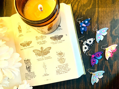 Shown next to an old book with drawings are six different colors of R+D 3D printed moth earrings. The six colors are Purple and Teal Wings, Pink Accents; Black and White Wings, Silver Accents; Purple and Navy Wings, Blue Accents; Hot Pink and Light Pink Wings, Yellow Accents; Mint and Light Blue Wings, White Accents; and White and Light Blue Wings, Gold Accents.