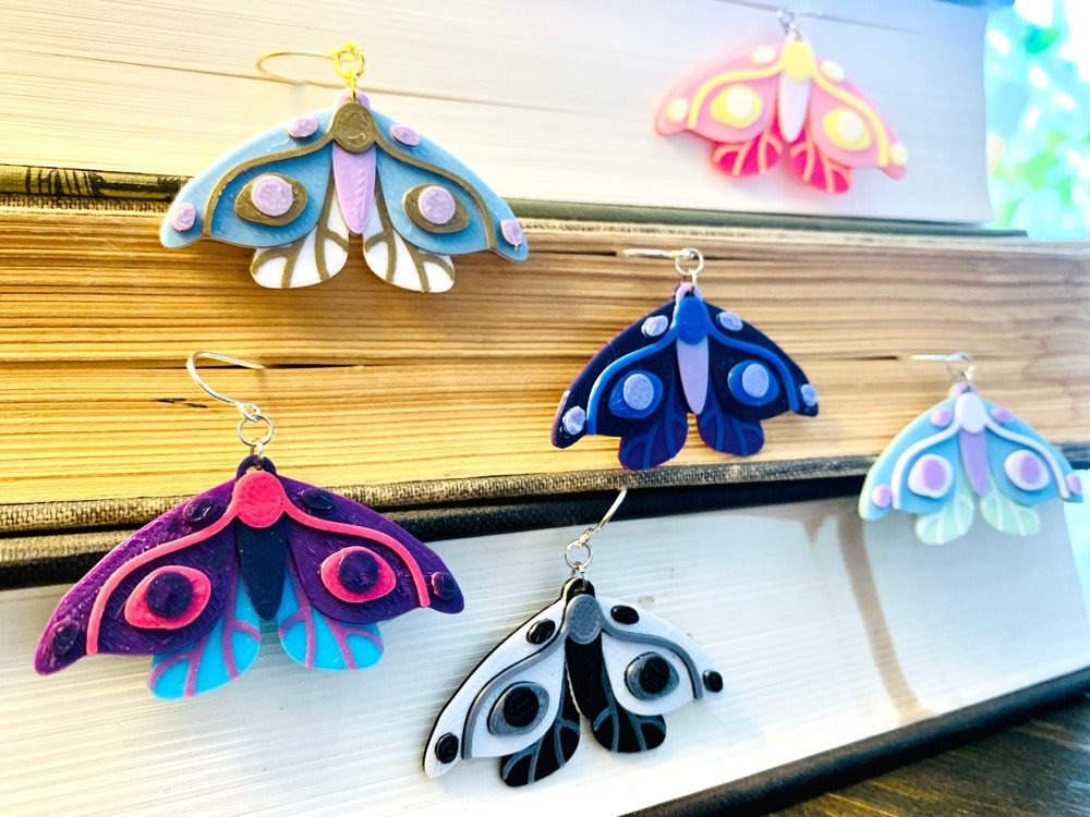Shown hanging out of the page edges of a stack of old books are six different colors of R+D 3D printed moth earrings. The six colors are Purple and Teal Wings, Pink Accents; Black and White Wings, Silver Accents; Purple and Navy Wings, Blue Accents; Hot Pink and Light Pink Wings, Yellow Accents; Mint and Light Blue Wings, White Accents; and White and Light Blue Wings, Gold Accents.