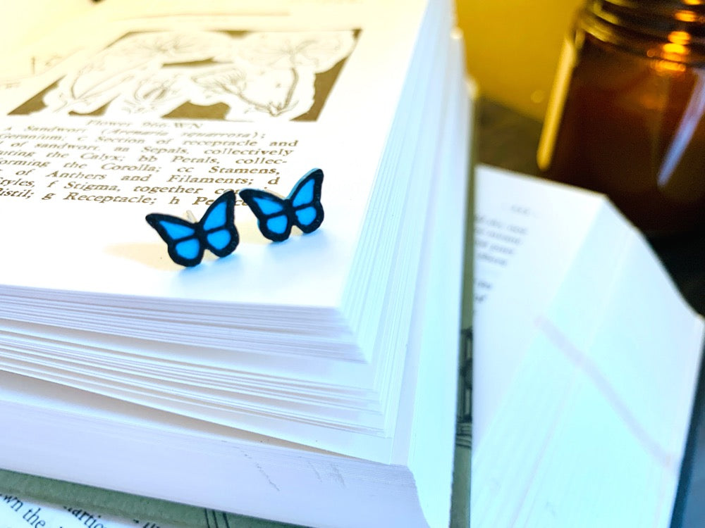 Resting on a stack of old books is a pair of R+D 3D Printed earrings. They are bright teal butterflies with a black outline. These studs are small and delicate. 