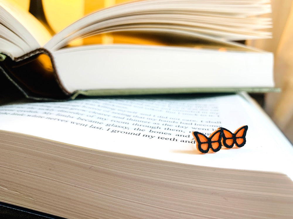 Resting on a stack of old books is a pair of R+D 3D Printed earrings. They are bright orange butterflies with a black outline. These studs are small and delicate.
