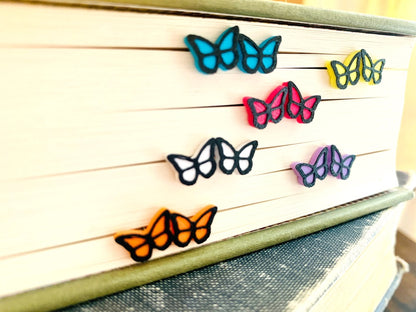 Sticking out of the edge of a book's pages are six pairs of R+D 3D Printed earrings. They are bright teal, white, orange, lavender purple, yellow, and hot pink butterflies with black outlines. These studs are small and delicate.