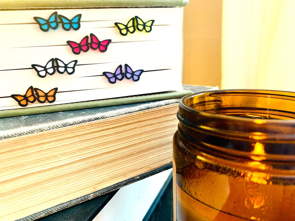 Sticking out of the edge of a book's pages are six pairs of R+D 3D Printed earrings. They are bright teal, white, orange, lavender purple, yellow, and hot pink butterflies with black outlines. These studs are small and delicate.