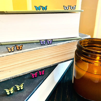 Resting on a stack of old books are six pairs of R+D 3D Printed earrings. They are bright teal, white, orange, lavender purple, yellow, and hot pink butterflies with black outlines. These studs are small and delicate.