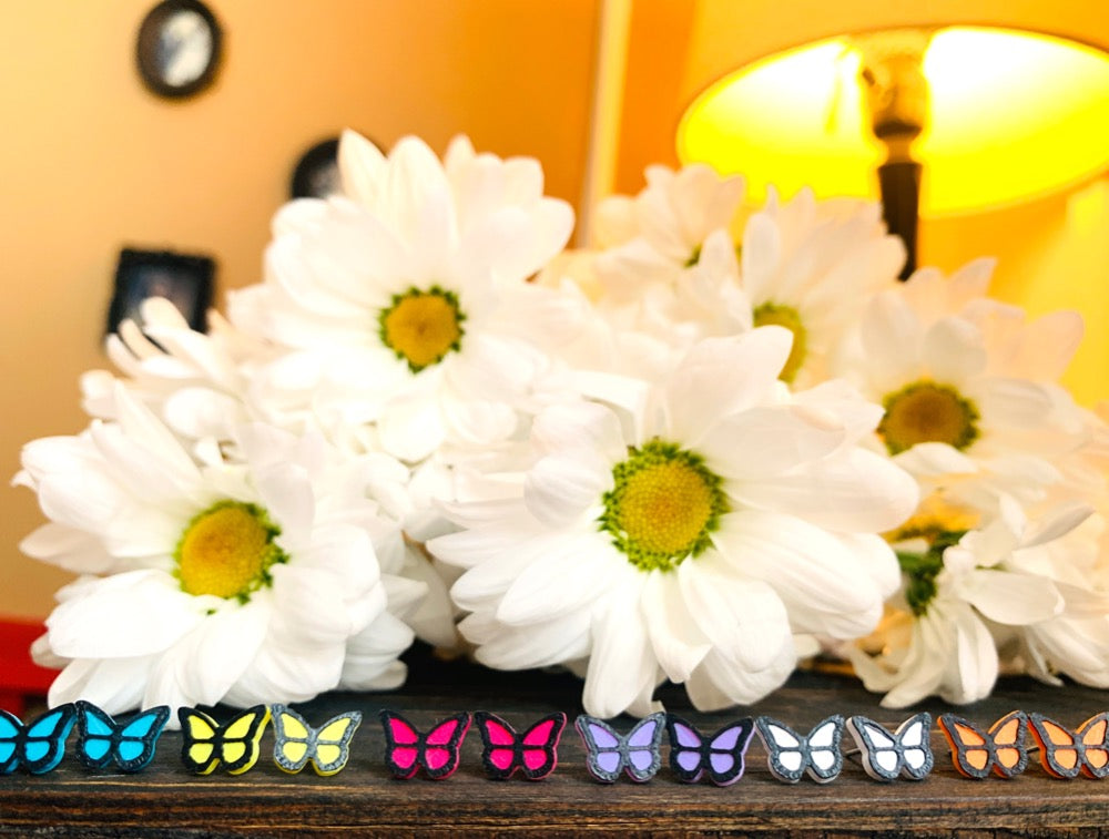 In front of a bouquet of daisy flowers is a line of R+D 3D printed earrings. They are small butterfly studs in six colors: teal, yellow, hot pink, purple, white, and orange. 