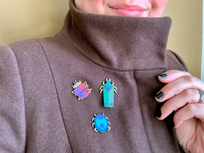 This is a close up of someone wearing a brown wool coat with a straight cowl neck. You can see a person smiling and holding up part of the coat to show a set of three R+D 3D printed pins. These three pins are each shaped as beetles with gold legs, rich colors of greens, merlot red, purples, and blues. 