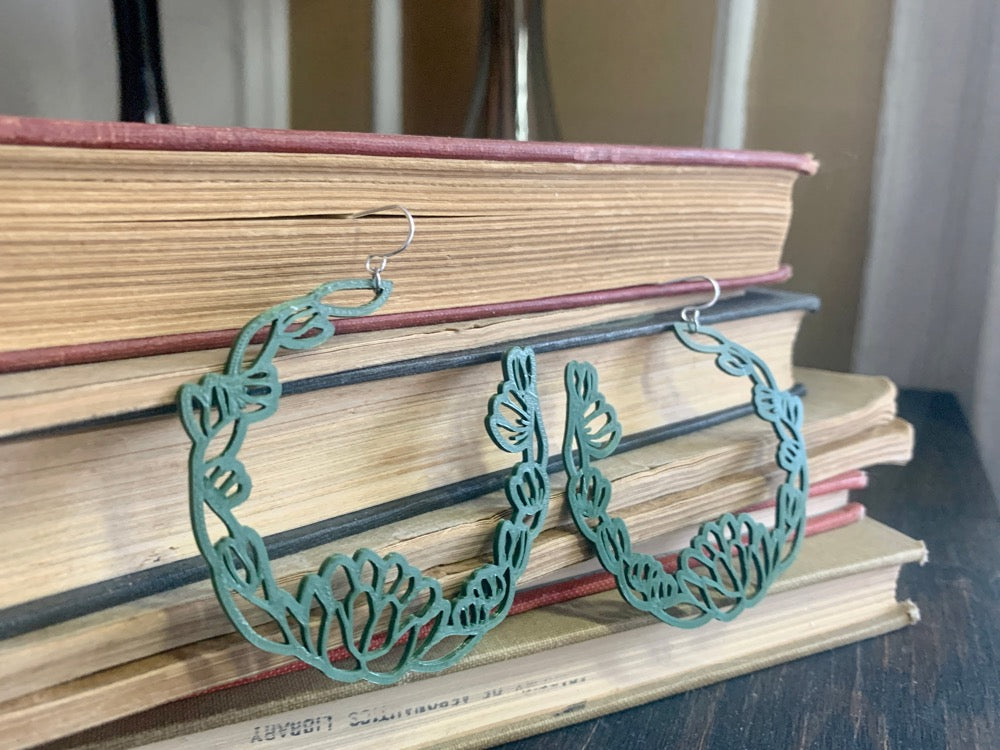 Resting in front of a stack of old yellowing books are two R+D floral hoop earrings. They are an olive green color with leaves and blooms twisting around to make a partial wreath shape.