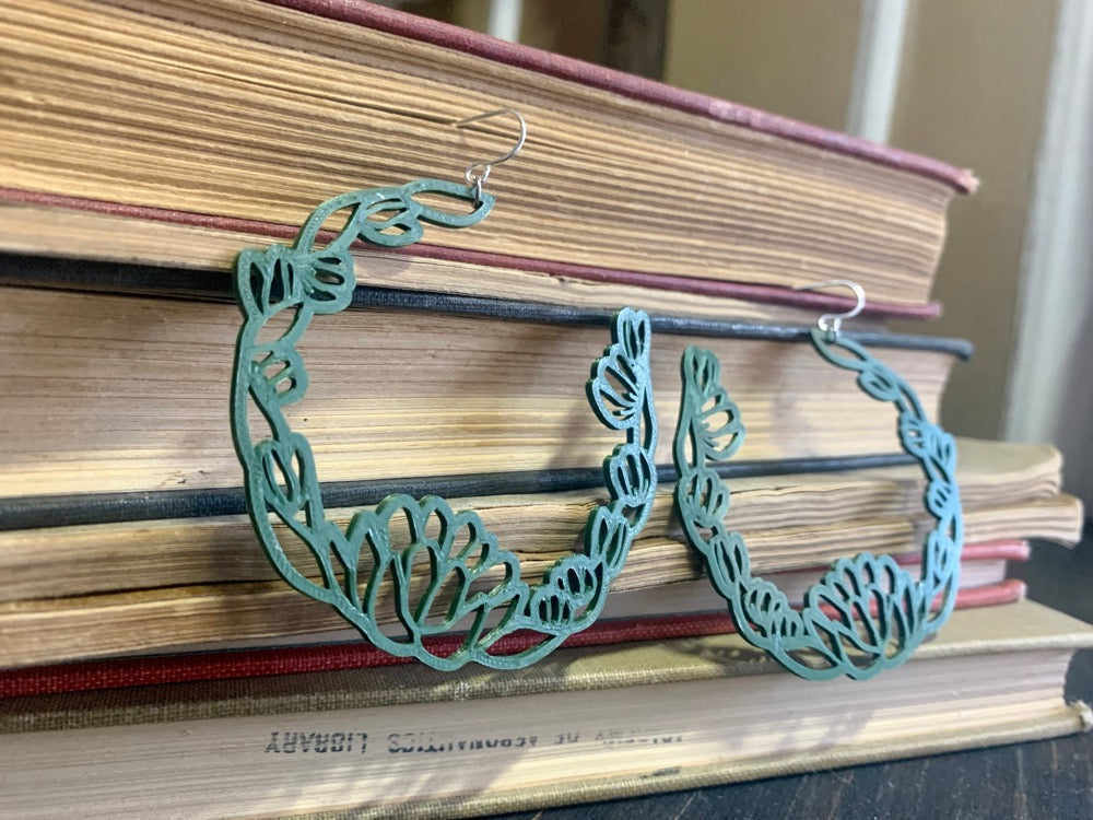 Resting in front of a stack of old yellowing books are two R+D floral hoop earrings. They are an olive green color with leaves and blooms twisting around to make a partial wreath shape.