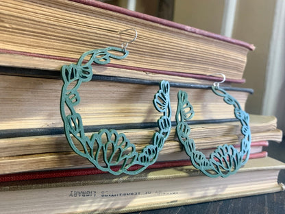 Resting in front of a stack of old yellowing books are two R+D floral hoop earrings. They are an olive green color with leaves and blooms twisting around to make a partial wreath shape.