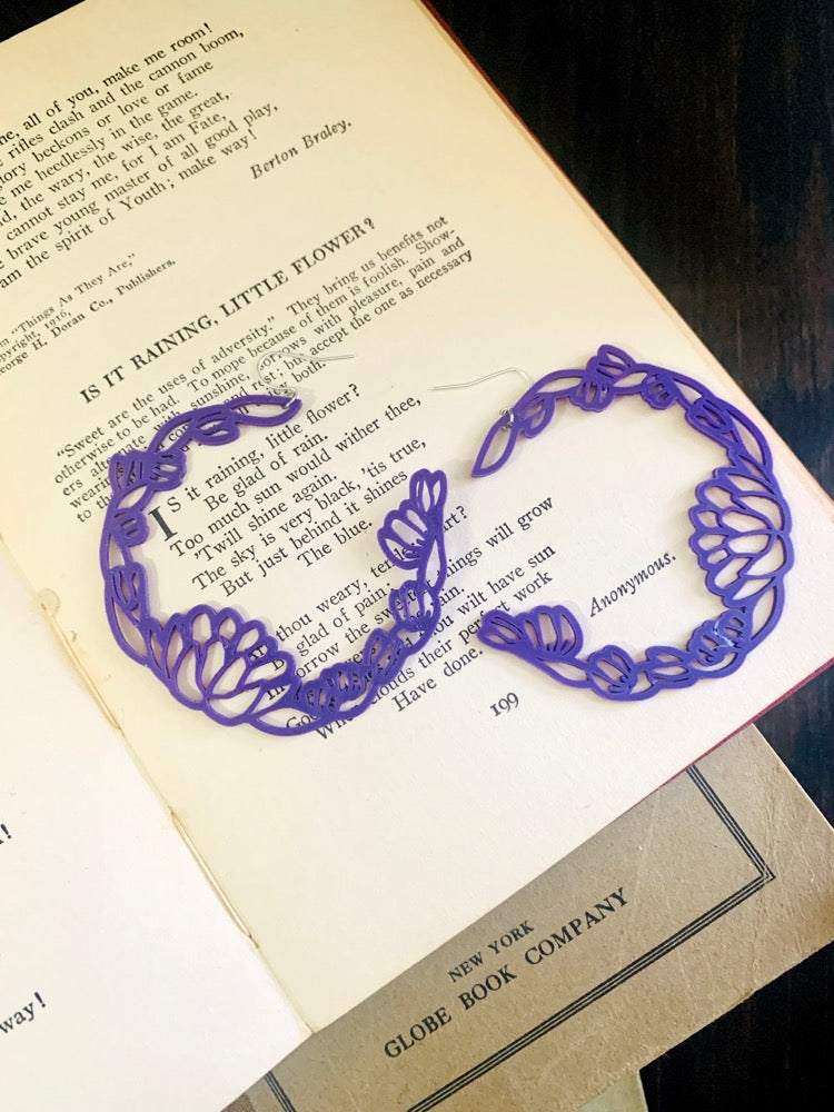 Resting inn open old book of poems are two R+D floral hoop earrings. They are a rich purple color with leaves and blooms twisting around to make a partial wreath shape.